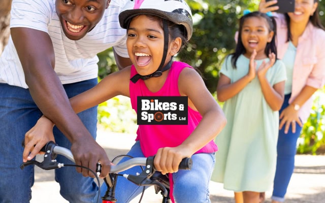 A child learning to ride a bicycle with help from an adult while family members watch