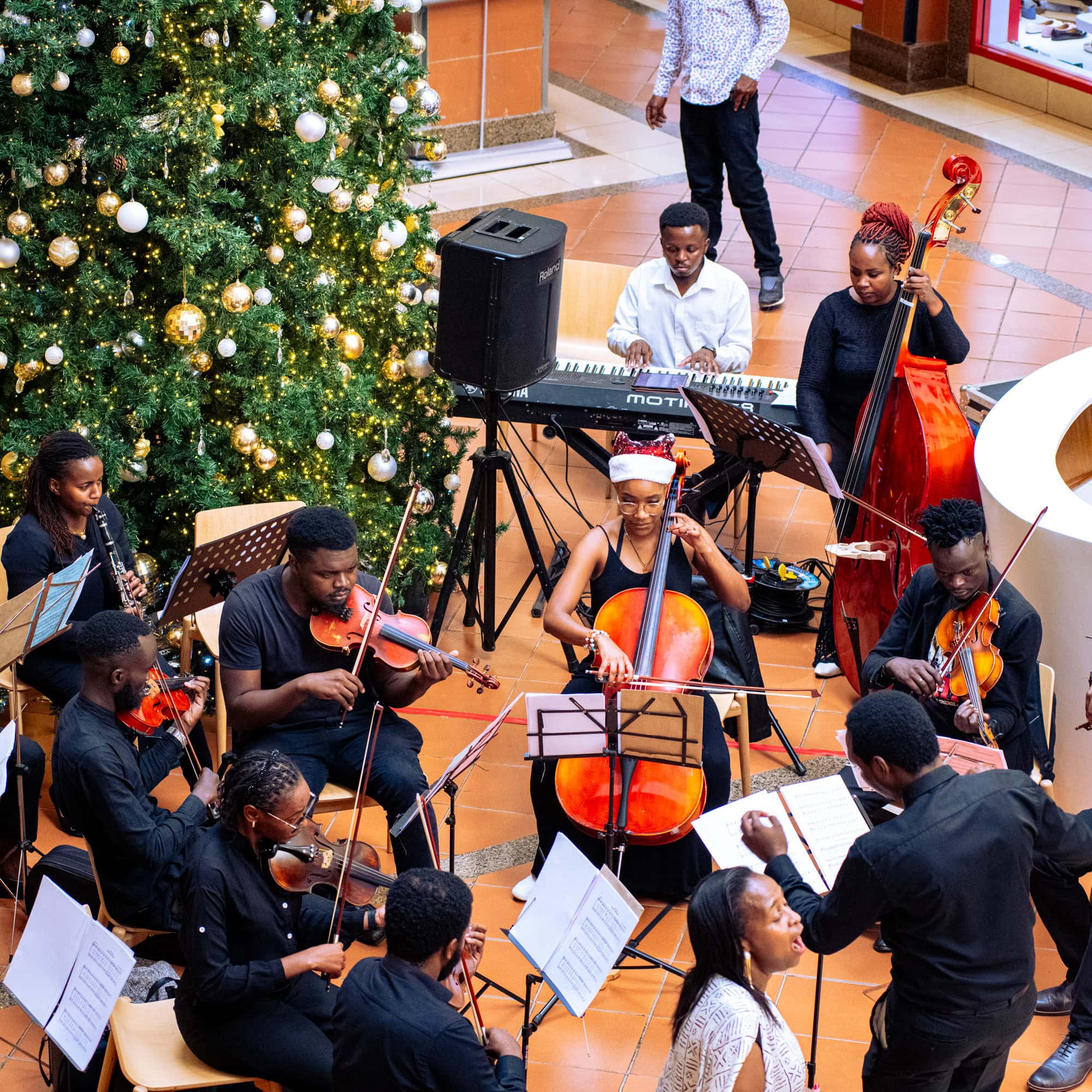 Musicians perform with strings and keyboard beside a decorated Christmas tree.