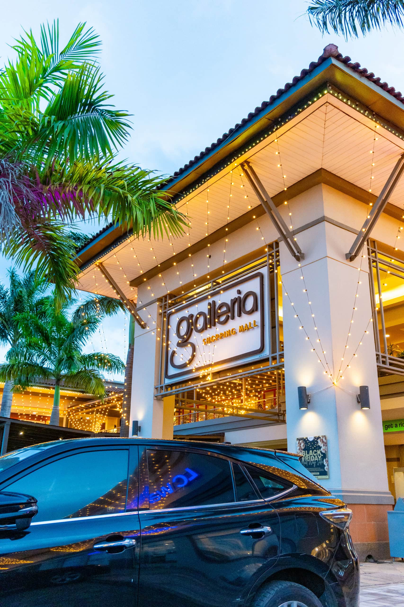 Galleria shopping mall exterior with string lights, palm trees, and a parked car