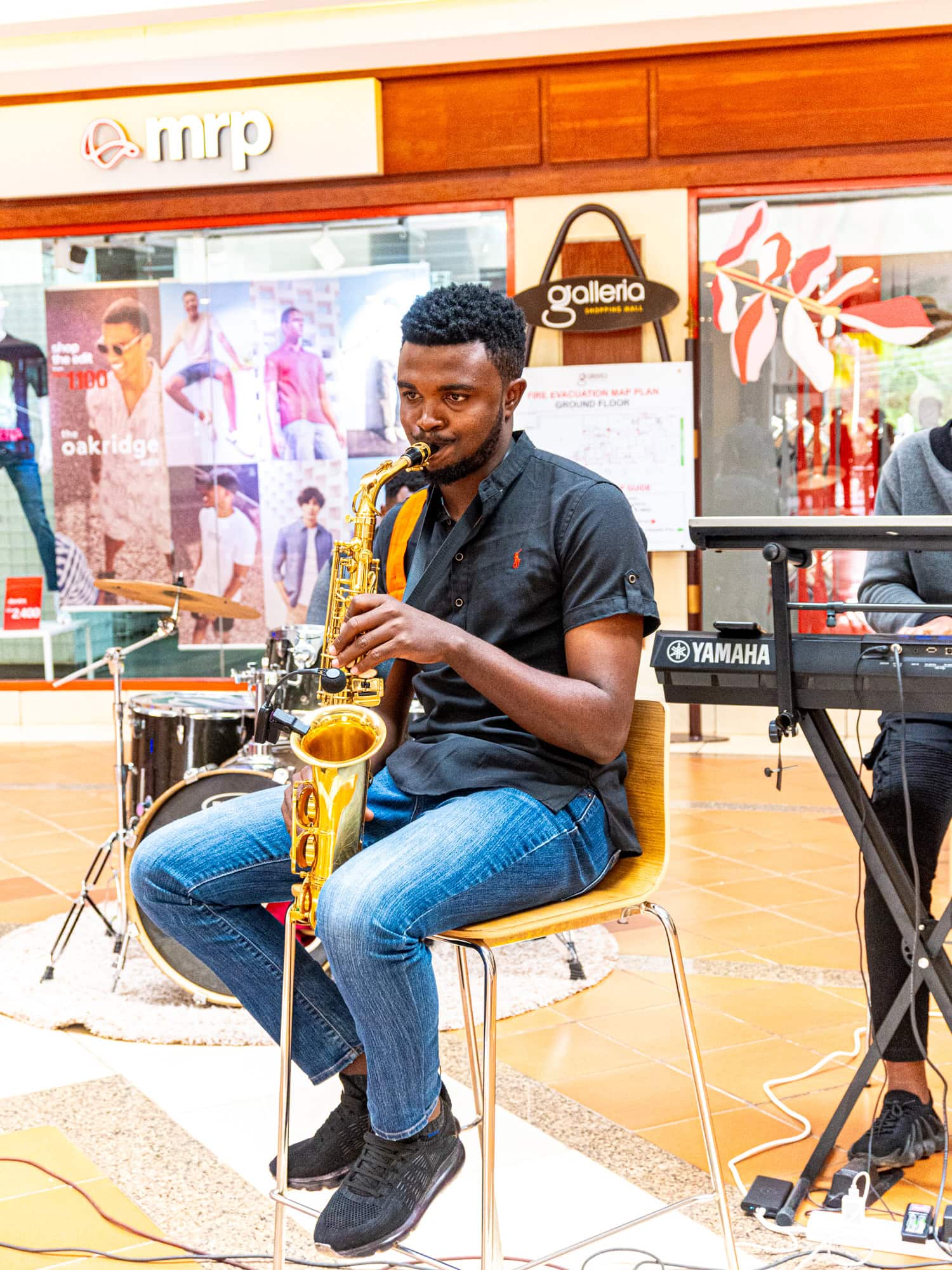 Musician seated and playing a saxophone beside a drum kit and Yamaha keyboard