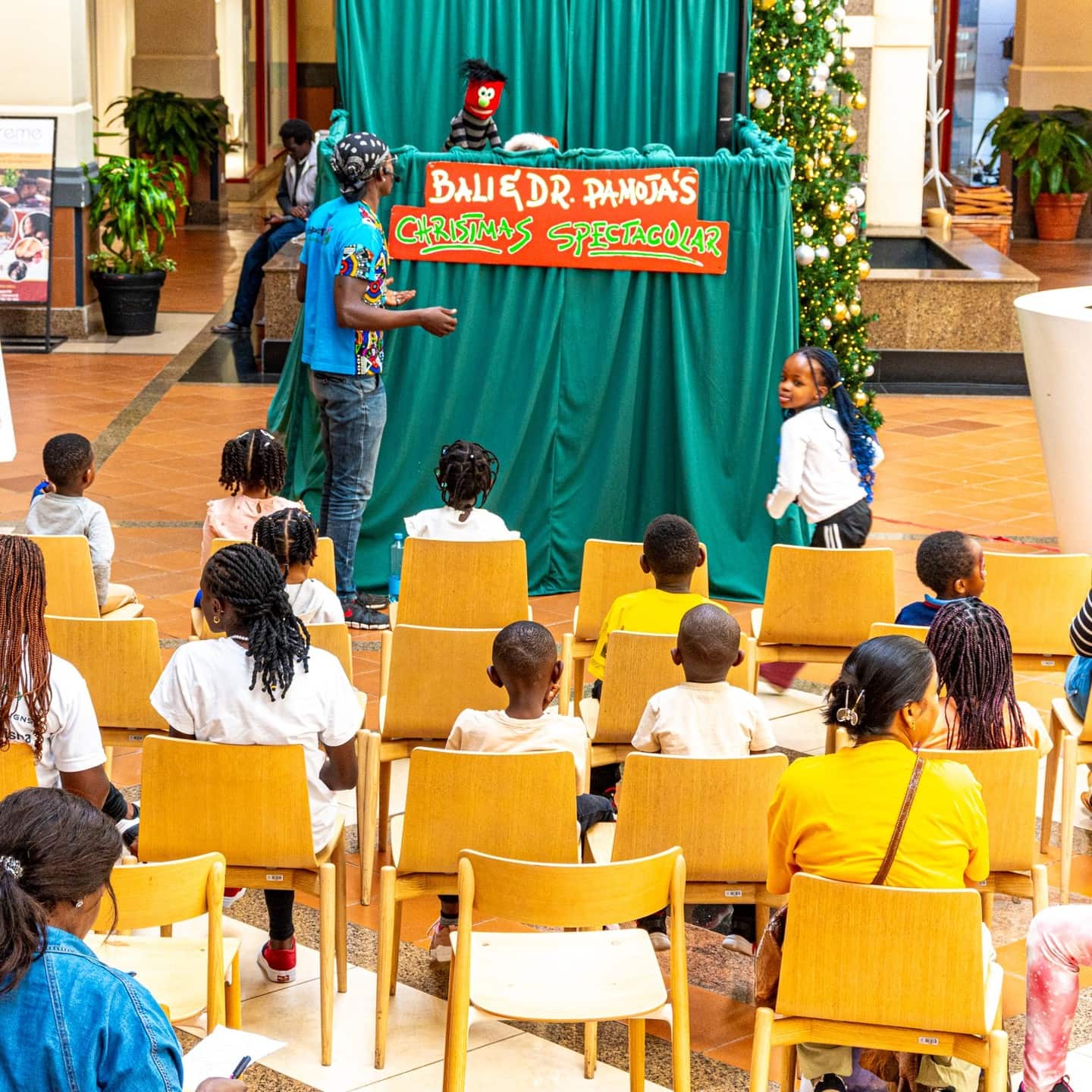 Children seated in rows watching a puppet show on a green stage in a shopping mall