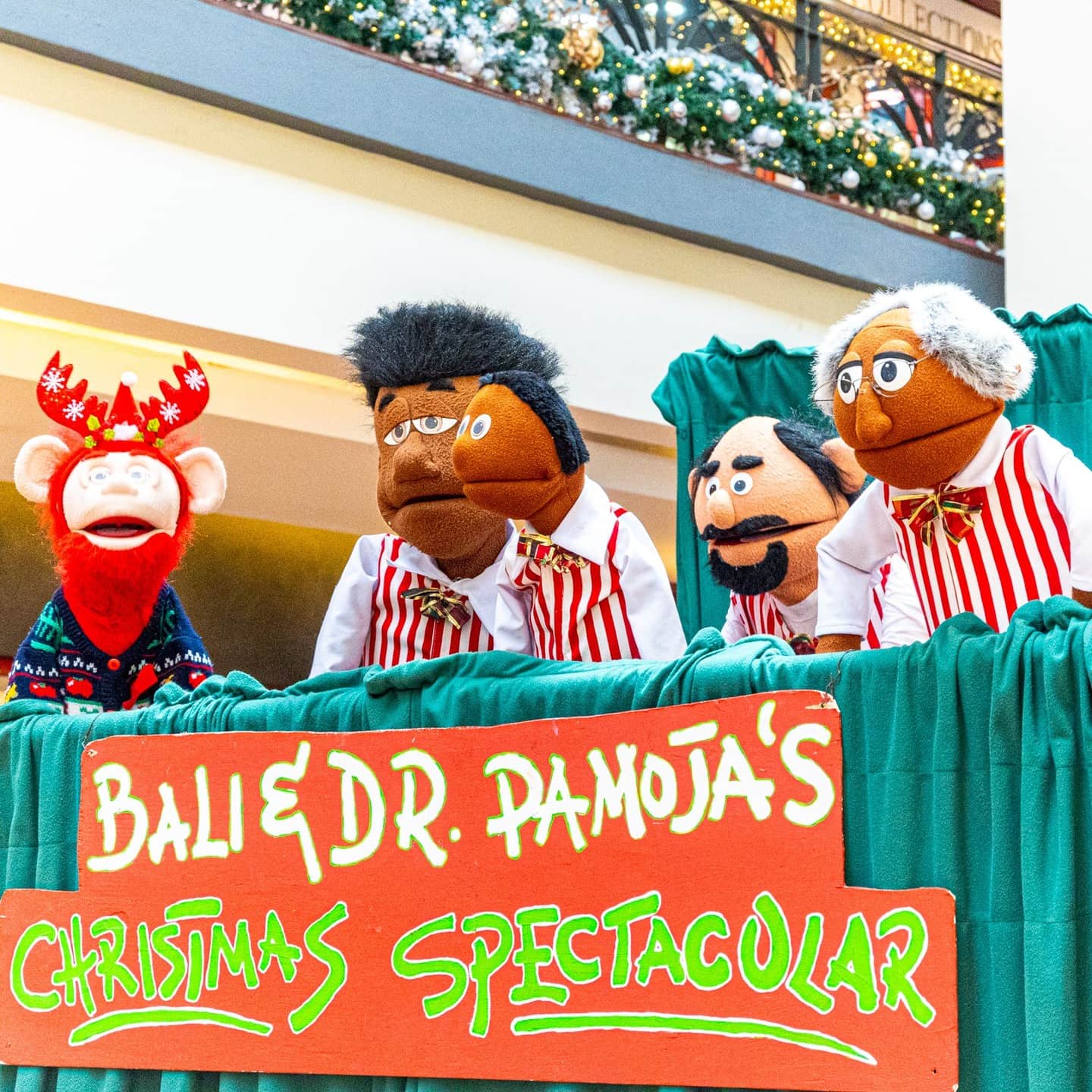 Four colorful puppets appearing above a green stage curtain, with a sign for a Christmas spectacular in front