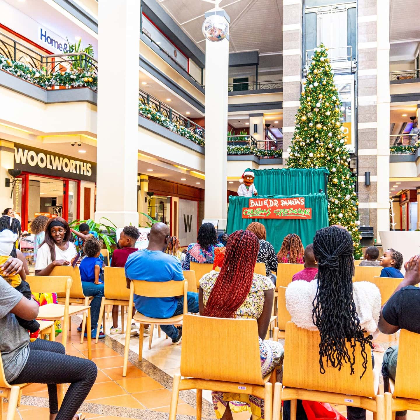 A seated audience watching a Christmas puppet show beneath a tall decorated Christmas tree in a shopping mall