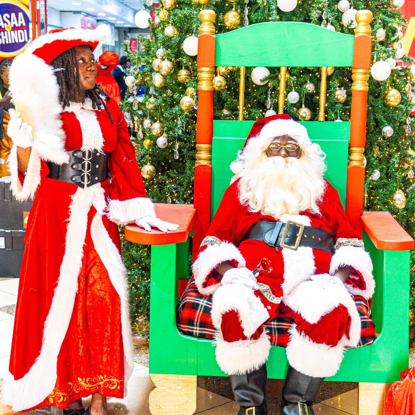 Santa sitting in a large green chair beside a woman in a red Santa-themed dress, with a decorated Christmas tree behind them