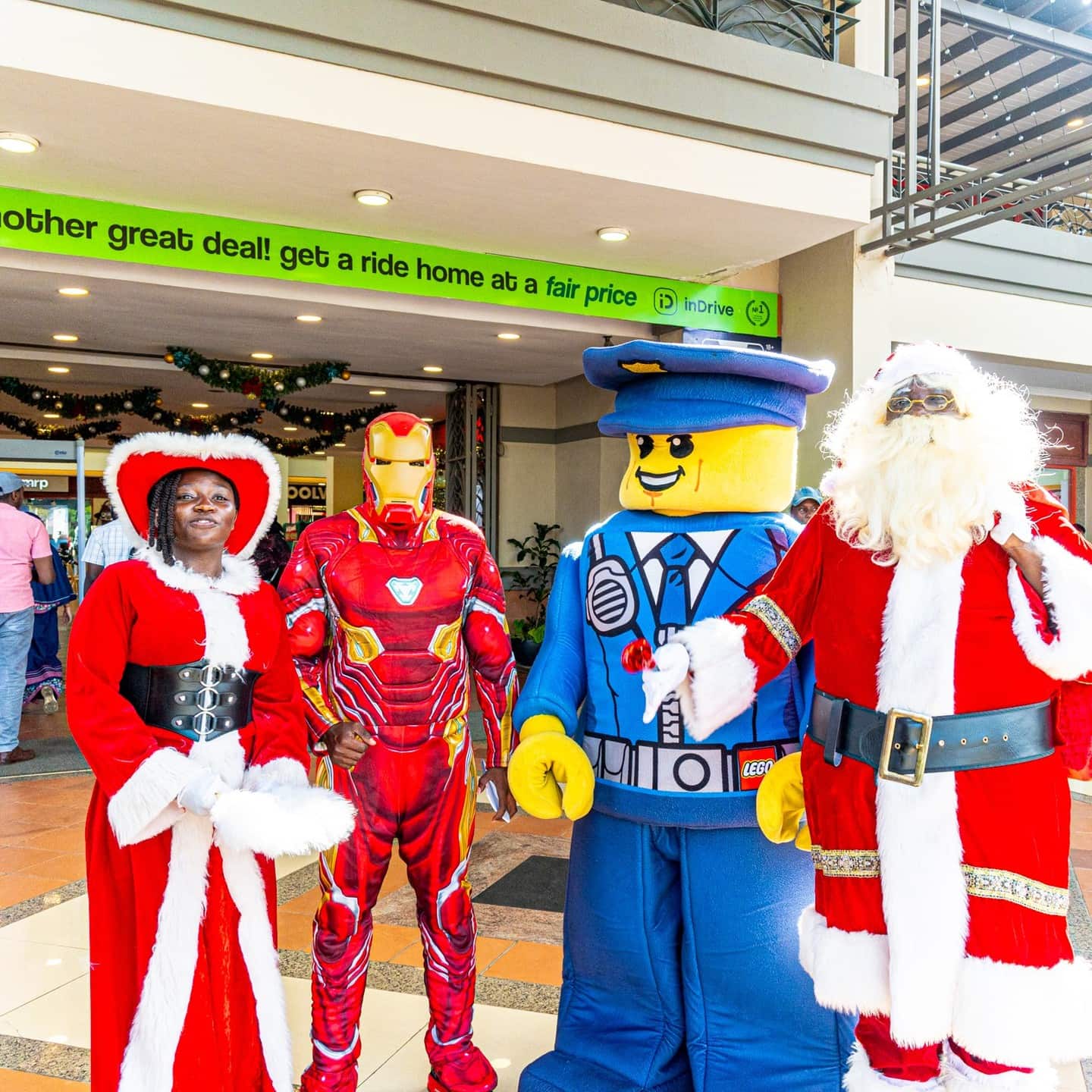 Santa, a woman in a red costume, an Iron Man character, and a blue-uniformed toy figure posing together in a shopping mall