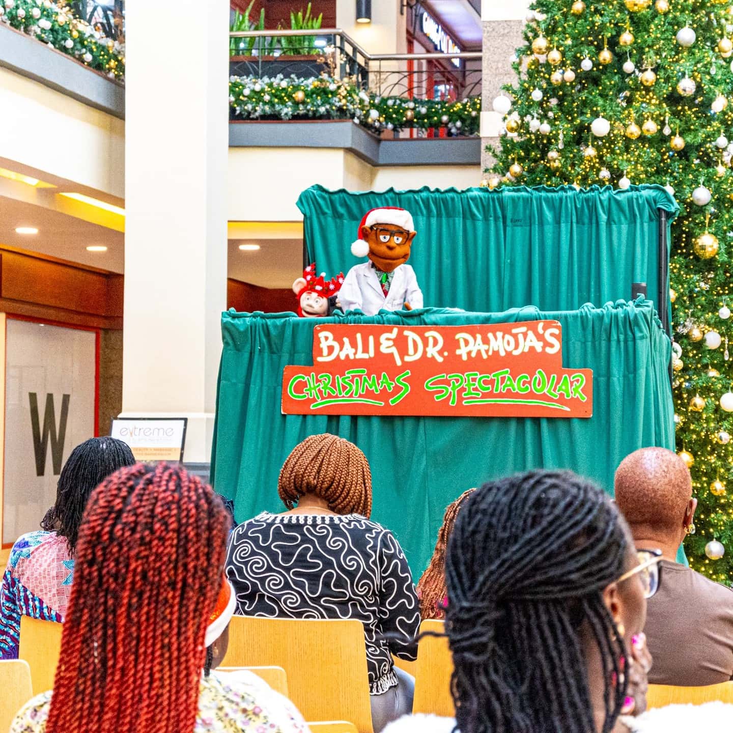 A puppet performer behind a green stage presenting a Christmas show to an audience in a decorated mall