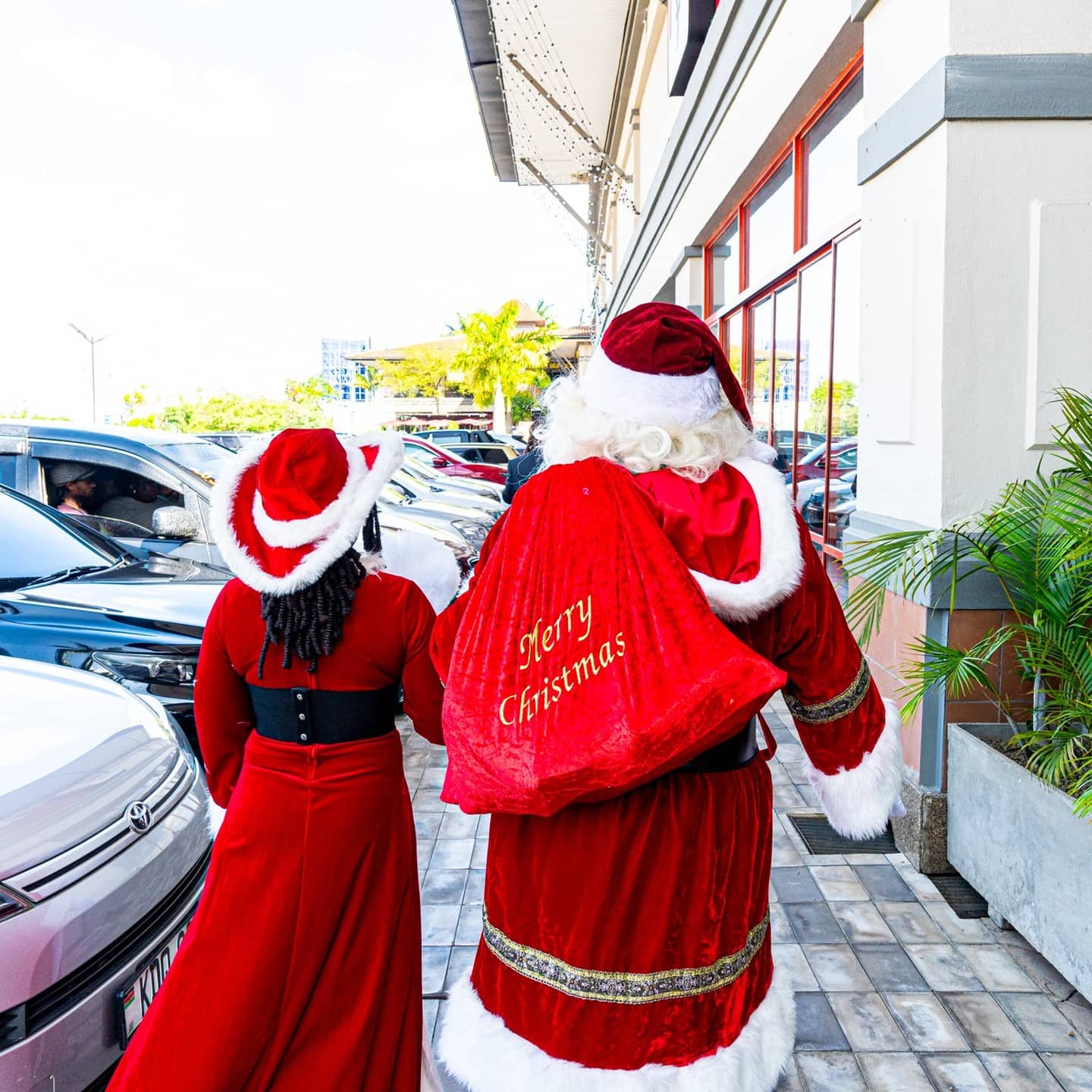 Santa and a companion walking beside parked cars outside a building, with Merry Christmas written on Santa's red cape