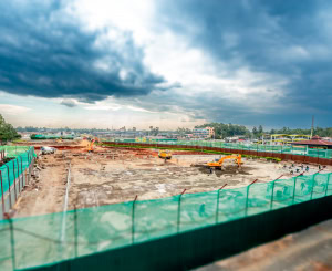 A broad construction site stretches beneath dramatic storm clouds, with machinery and green safety fencing visible.