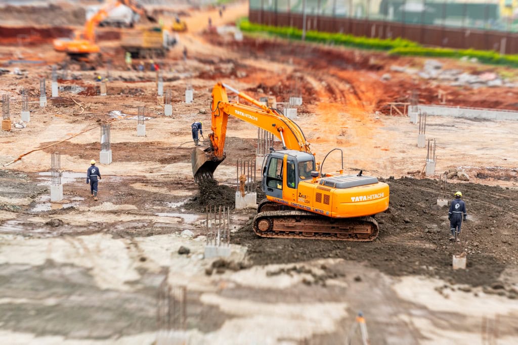 An orange excavator lifts soil from a trench while workers and reinforced foundation columns surround it.