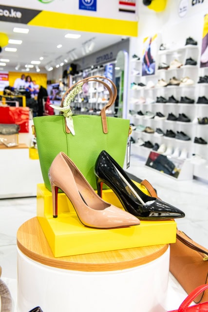 Green handbag displayed beside nude and black pointed heels on a yellow pedestal.