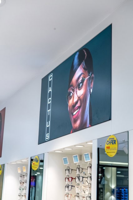 Eyewear store interior showing wall-mounted frames, offer signs, and a large portrait advertisement