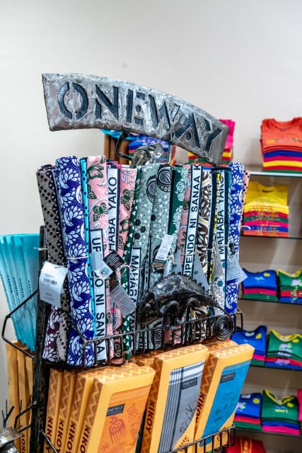 Rack of patterned fabric accessories beneath a metal ONE WAY sign, with colorful clothing stacked behind it