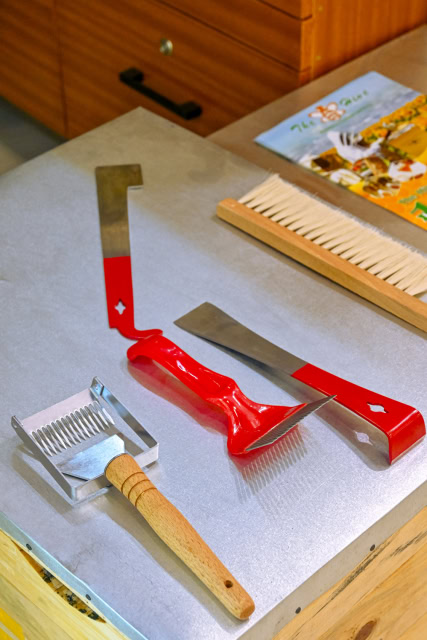 Red-handled beekeeping tools, a wooden brush, and a metal uncapping tool arranged on a tabletop.