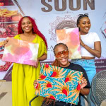 Three participants display colourful fabric artwork at the Frame & Fabric Workshop, with scissors and materials visible on the table.