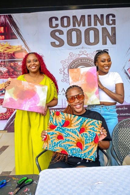 Three participants display colourful fabric artwork at the Frame & Fabric Workshop, with scissors and materials visible on the table.