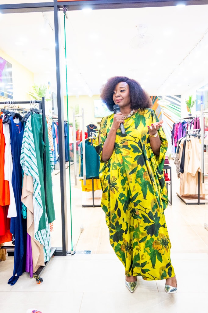 Connie Oluoch speaks into a microphone while standing among clothing racks at Style, Shop & Sip.