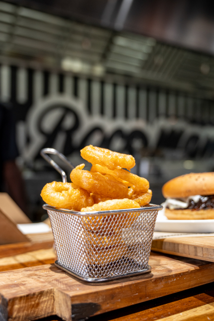Golden fried onion rings served in a small metal basket with a burger in the background.