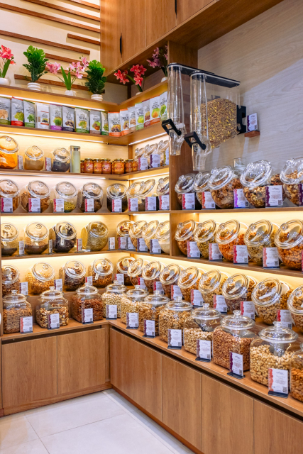 Corner of a retail store showing labelled glass jars, packaged foods and dispensers on illuminated wooden shelves.