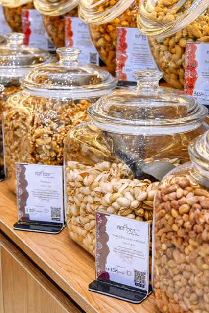 Glass jars of pale seeds and seasoned nuts displayed with product cards on a wooden shelf.