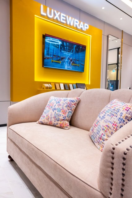 Cream sofa with patterned cushions in front of a yellow LUXEWRAP showroom display wall