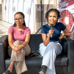 Women participating in a wellness discussion Two women sit on a lounge sofa during a wellness discussion, with one holding a microphone