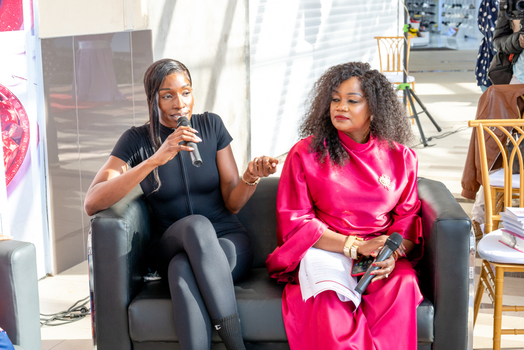 A speaker gestures while holding a microphone during a discussion with another woman seated beside her.