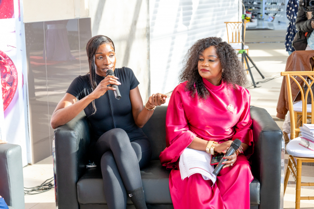 A speaker gestures while holding a microphone during a discussion with another woman seated beside her.