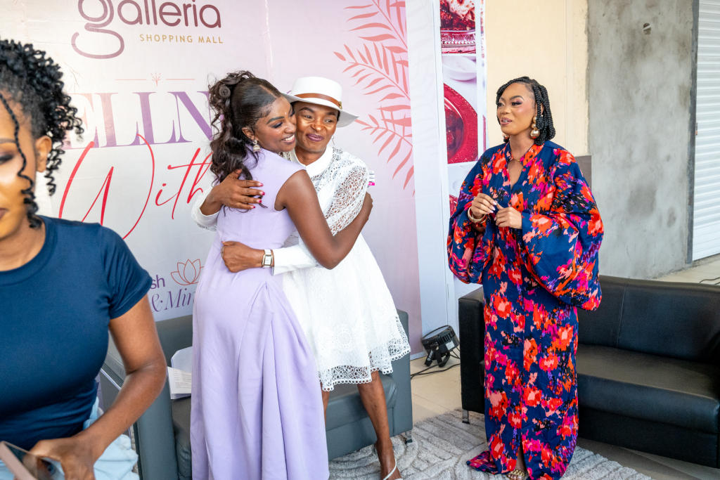 Two women embrace in front of an event backdrop while a woman in a floral outfit looks on.