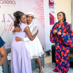 Celebratory Embrace at Wellness Within Event Two women embrace in front of an event backdrop while a woman in a floral outfit looks on.
