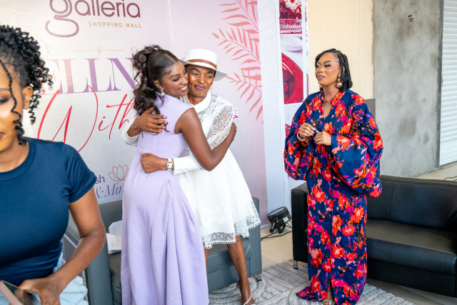 Two women embrace in front of an event backdrop while a woman in a floral outfit looks on.