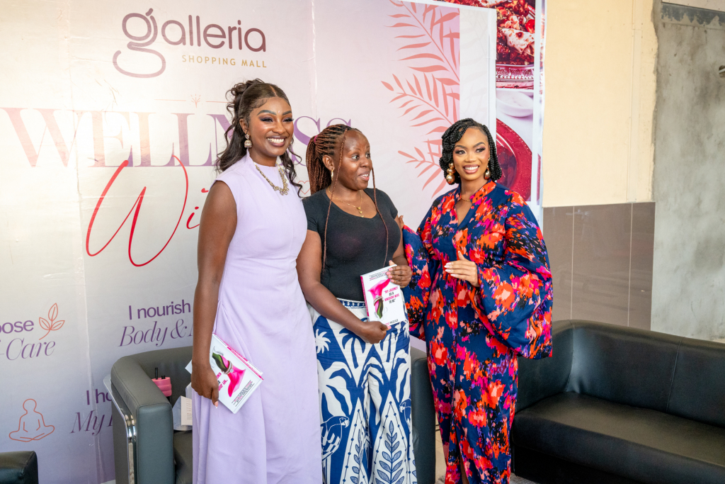 Three smiling women pose together in front of a Wellness Within backdrop at Galleria Shopping Mall.