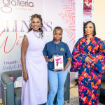 Three Women at the Wellness Within Backdrop Three women pose together in front of a Galleria Shopping Mall Wellness Within backdrop, with the central attendee holding a booklet.