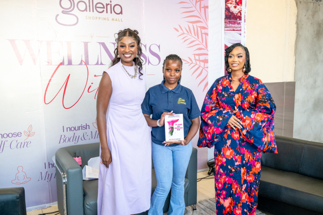 Three women pose together in front of a Galleria Shopping Mall Wellness Within backdrop, with the central attendee holding a booklet.