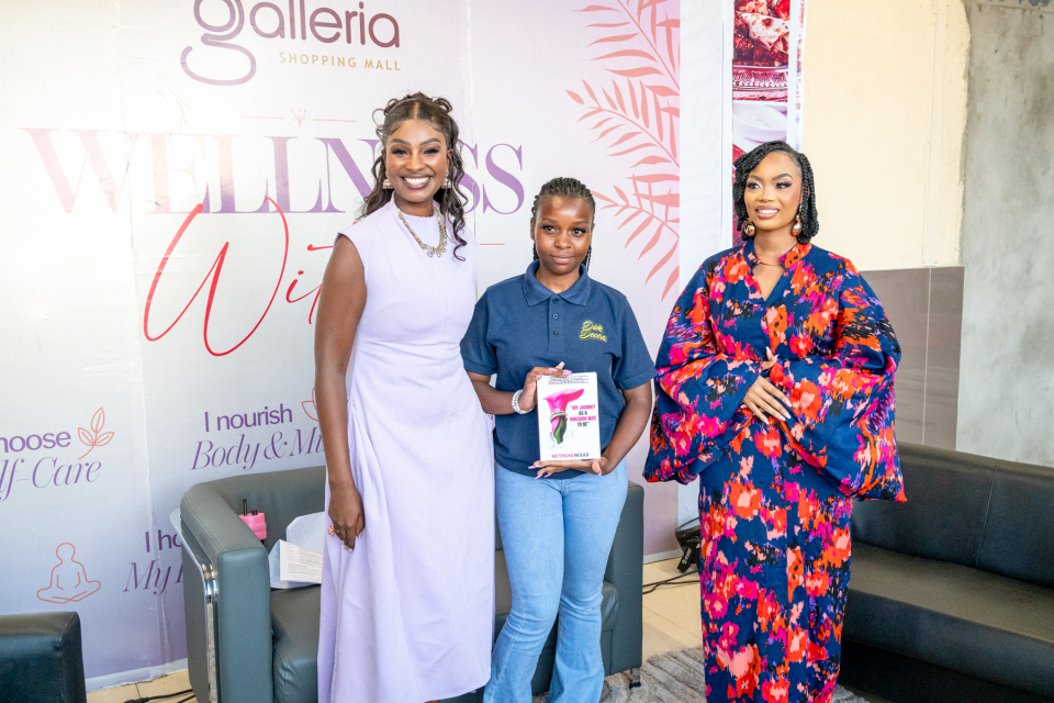 Three Women at the Wellness Within Backdrop Three women pose together in front of a Galleria Shopping Mall Wellness Within backdrop, with the central attendee holding a booklet.