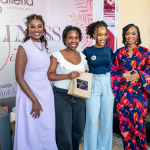 Four women pose together at the wellness event Four women smile for a group photo, with two holding a small gift bag in front of a Galleria backdrop.