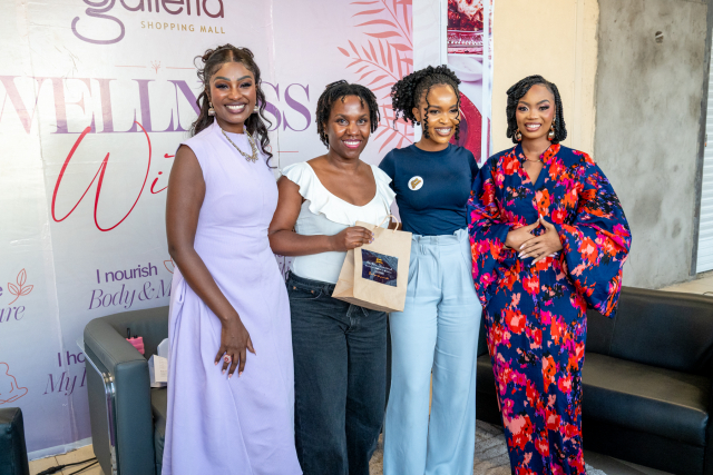 Four women smile for a group photo, with two holding a small gift bag in front of a Galleria backdrop.