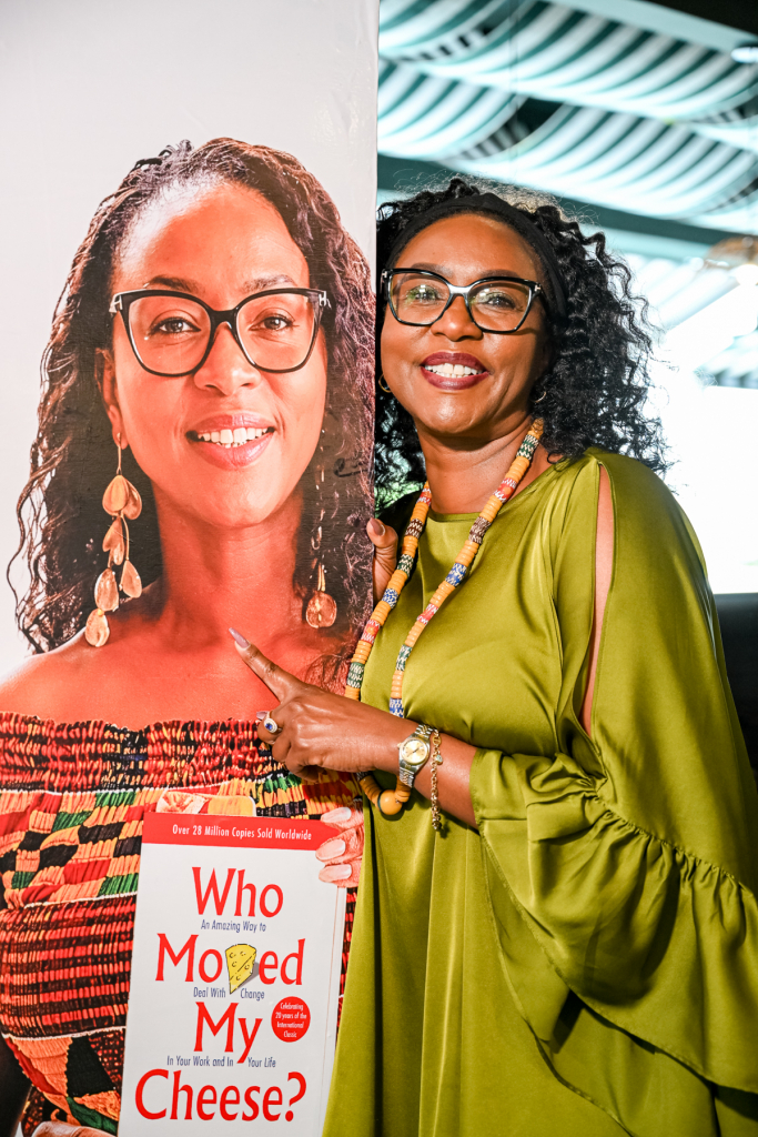 Smiling woman in green poses beside a large portrait display showing a woman holding the book Who Moved My Cheese?