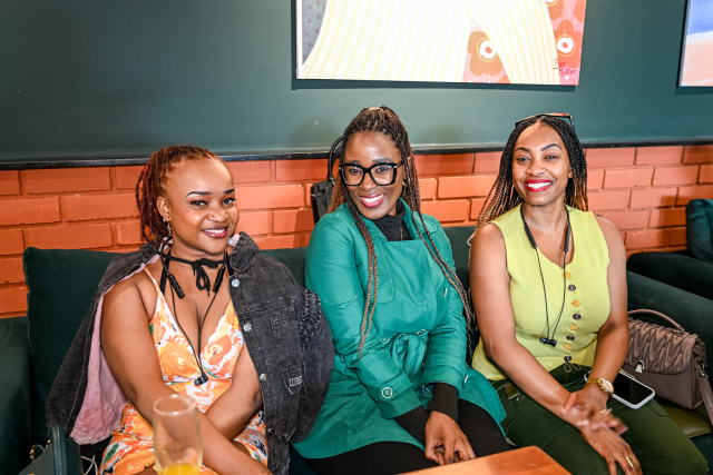 Three smiling women sit together on green lounge chairs in a mall event space.