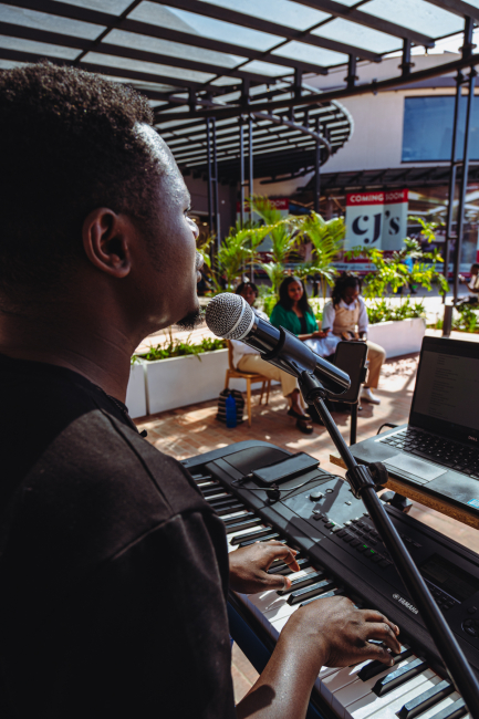 Keyboard player performing outdoors with a microphone and laptop as people sit nearby.