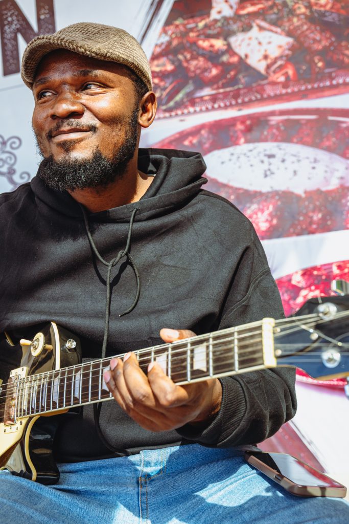 A smiling guitarist in a flat cap plays a gold electric guitar