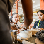 Participants work together with clay around a large container at a workshop table.
