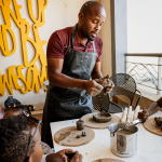 An instructor demonstrates clay work to children gathered around a workshop table.