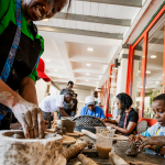 Smiling participant shapes clay at a pottery workshop while other people work in the background.