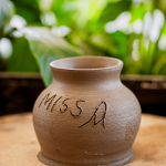 A handmade clay vessel with handwritten markings sits on a pottery work surface.