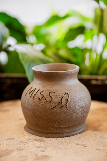 A handmade clay vessel with handwritten markings sits on a pottery work surface.