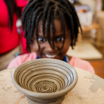 A smiling child looks over a shallow patterned clay bowl on a worktable.