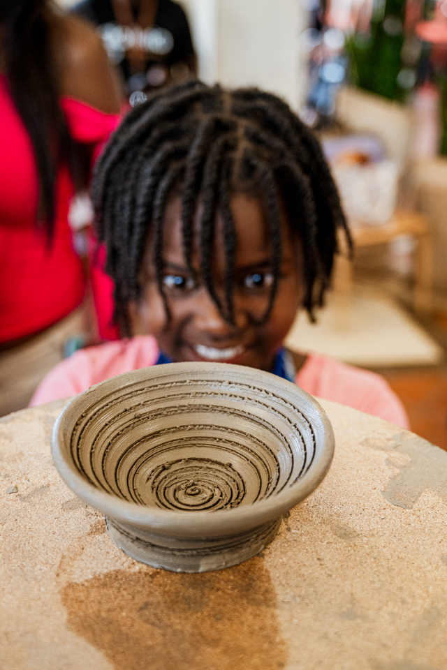 A smiling child looks over a shallow patterned clay bowl on a worktable.