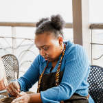 Participant in a blue sweater shapes a clay vessel by hand at a pottery table.