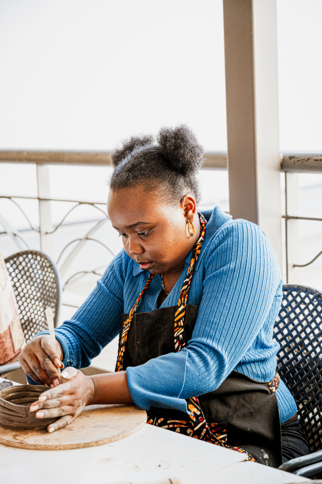 Participant in a blue sweater shapes a clay vessel by hand at a pottery table.