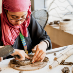 A participant in a pink headscarf shapes clay on a pottery work board.
